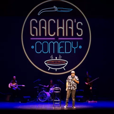 Escenario con monologuista y banda musical al fondo bajo iluminación azul.