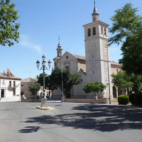 Plaza con iglesia de torre alta, farolas y edificio institucional entre árboles