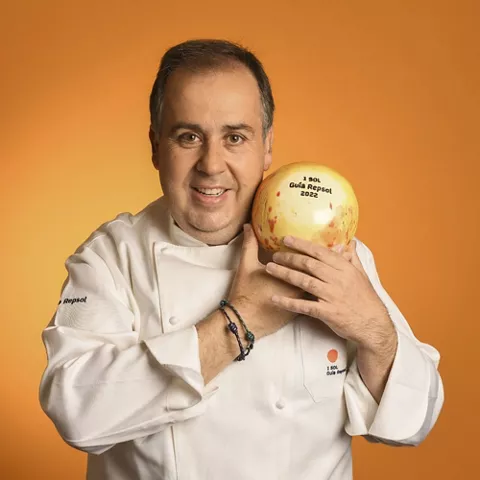 Cocinero sonriente sostiene un galardón redondo sobre fondo naranja.