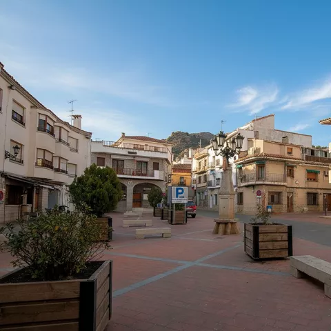Una plaza de pueblo empedrada con maceteros de madera, bancos y edificios residenciales de varios pisos bajo un cielo azul despejado.