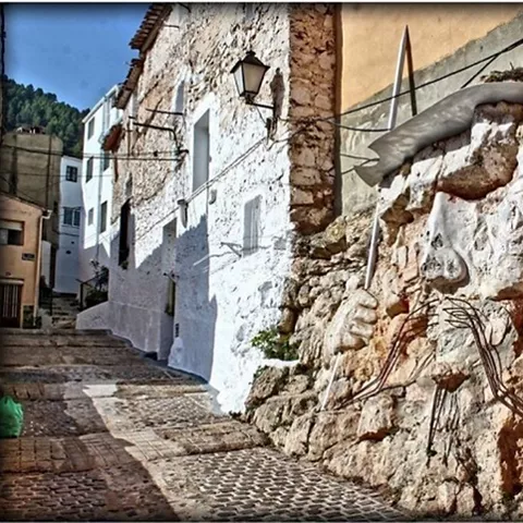 Calle empedrada con casas blancas y escultura integrada en un muro de piedra.