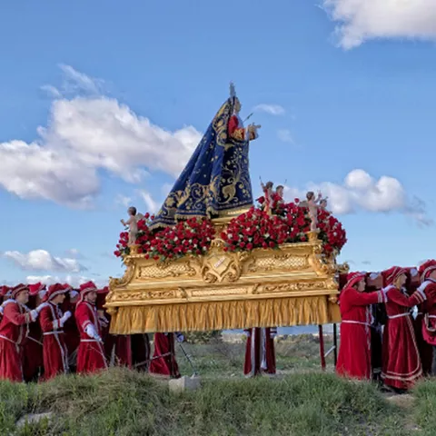 Imagen religiosa sobre un trono dorado, portada por personas vestidas de rojo en un entorno abierto.