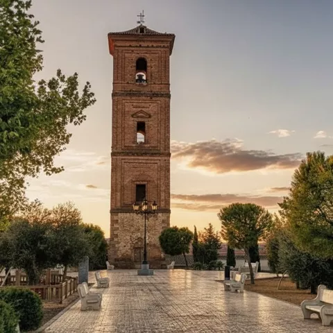 Plaza ajardinada con torre de ladrillo al fondo y bancos de piedra.