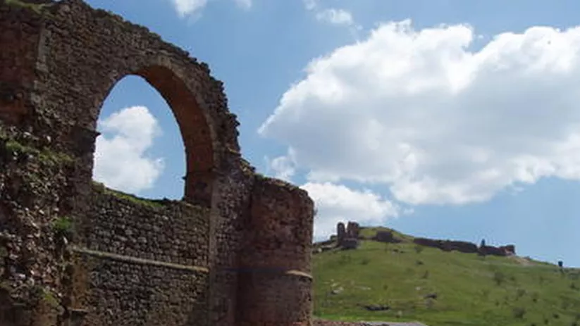 Estructura de piedra con arco conservado en ladera verde y cielo con nubes.