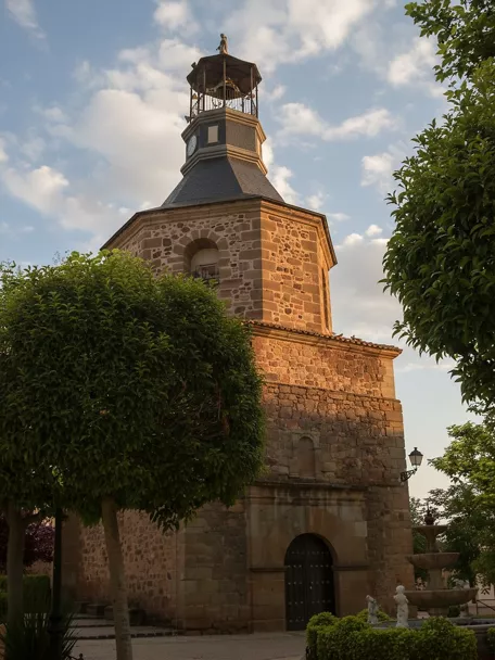 Torre de la iglesia rodeada de vegetación y fuente en plaza