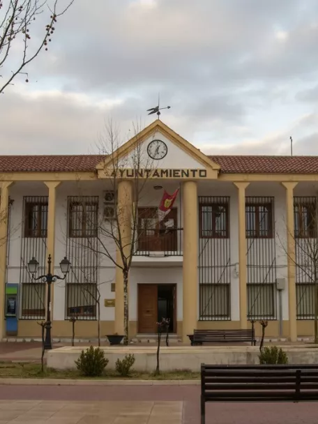 Fachada del Ayuntamiento de Arenas de San Juan con columnas, reloj central y plaza con bancos en primer plano.
