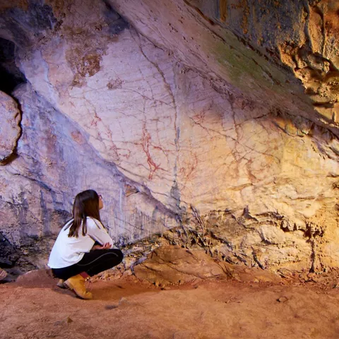 Persona agachada observando pinturas antiguas en la pared de una cueva.
