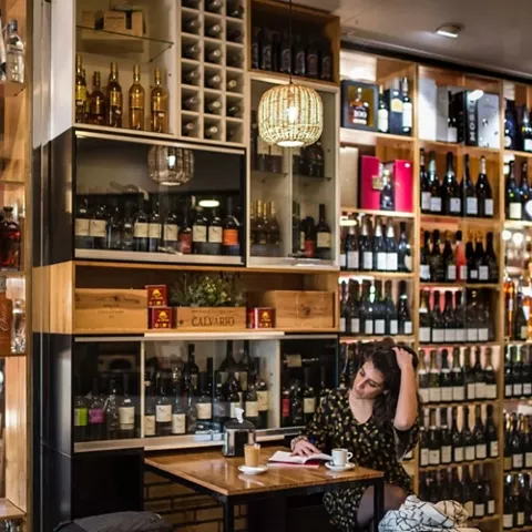 Interior de tienda de vinos con estanterías llenas y clienta sentada.