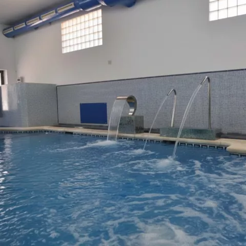 Piscina interior con cascada y chorros en fila, agua burbujeante y ventanales altos.