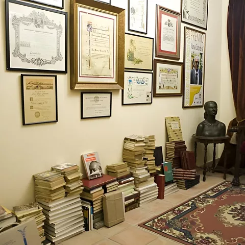 Despacho antiguo con libros apilados y diplomas en la pared.