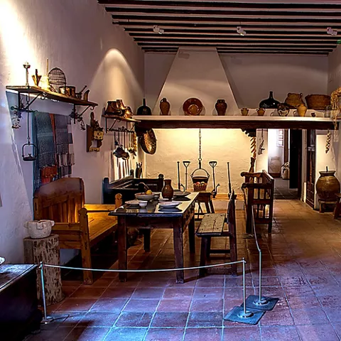 Cocina tradicional con chimenea, mesa larga y utensilios colgados en la pared.