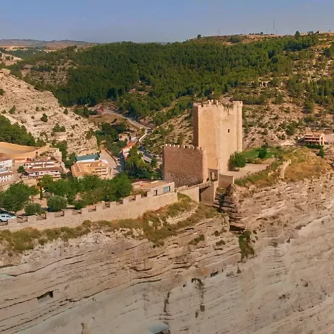 Vista aérea de una fortaleza de piedra sobre un acantilado, rodeada de bosque y casas en el valle.
