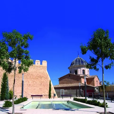 Plaza ajardinada con fuente frente a muralla y edificio con cúpula.