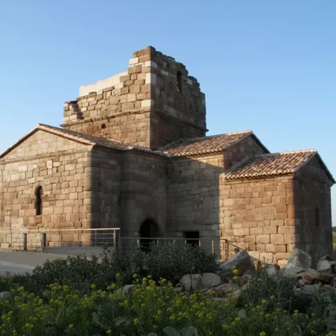 Vista lateral de la iglesia de piedra con torre, rodeada de vegetación baja y rocas.