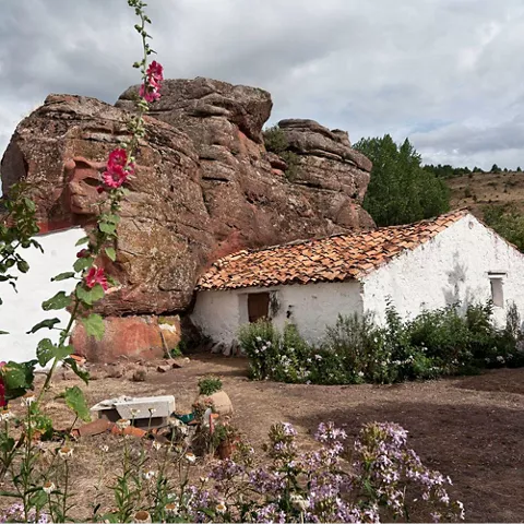 Casa rural de paredes blancas integrada en grandes rocas, rodeada de flores silvestres.