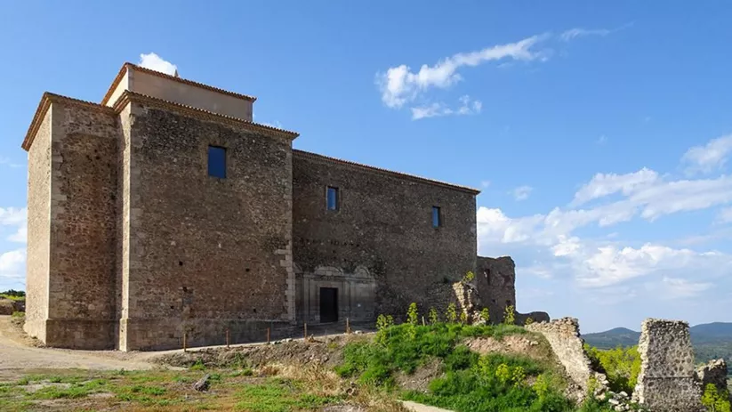 Edificio histórico de piedra sobre una colina, junto a restos de muralla y paisaje natural.