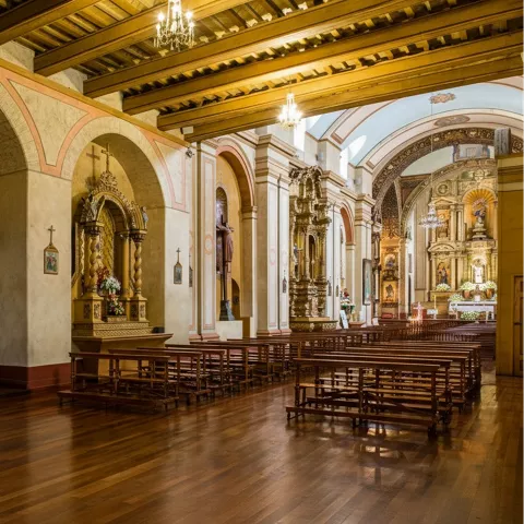 Nave de iglesia con bancos de madera y altares laterales ornamentados.