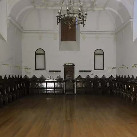Sala amplia con suelo de madera, sillería oscura tallada y lámpara colgante bajo bóveda blanca.