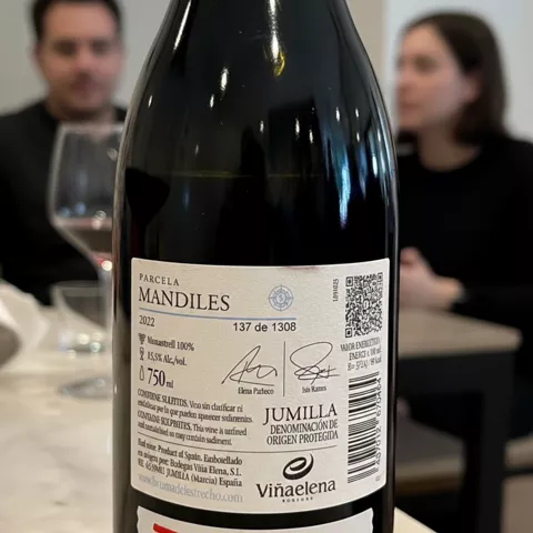 Primer plano de etiqueta trasera de una botella de vino sobre mesa, con comensales desenfocados.