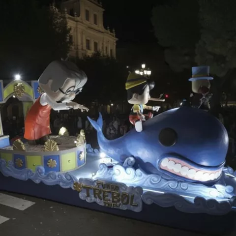Carroza iluminada con figuras infantiles sobre una ballena azul durante desfile nocturno.