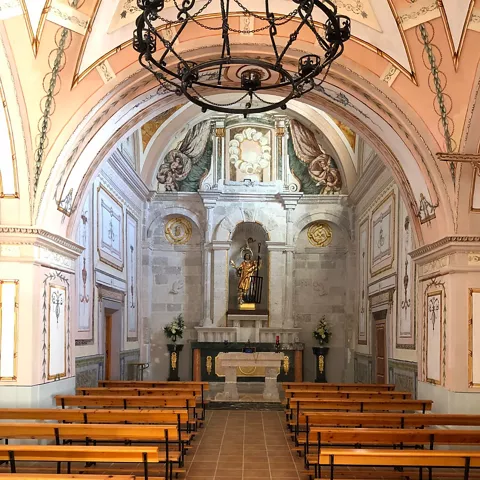 Interior de ermita vacía con altar decorado, bancos de madera y bóveda pintada.