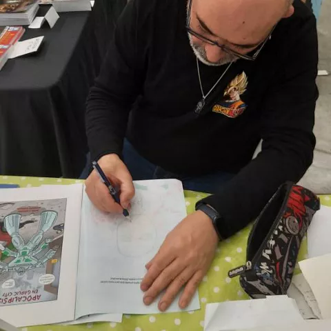 Ilustrador firmando y dibujando en mesa con cómics expuestos.