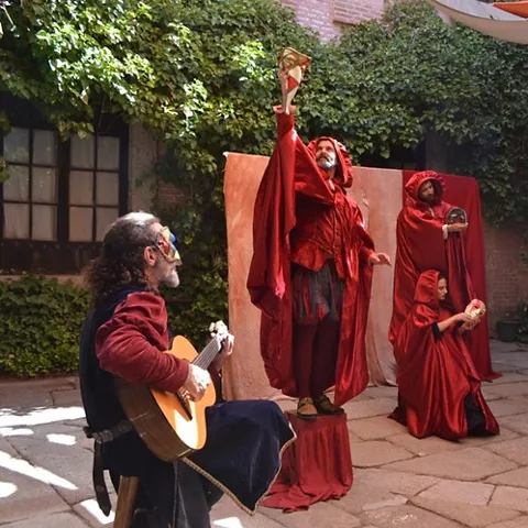 Representación teatral al aire libre con actores vestidos con capas rojas y músico tocando la guitarra en un patio con vegetación.