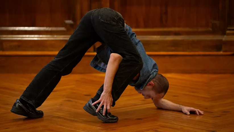 Bailarín en el suelo haciendo una postura acrobática en una sala con suelo de madera.