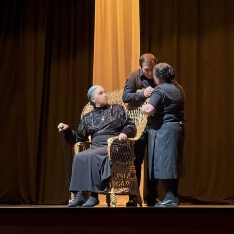 Escena teatral con tres personas alrededor de una silla de mimbre bajo cortinas doradas.