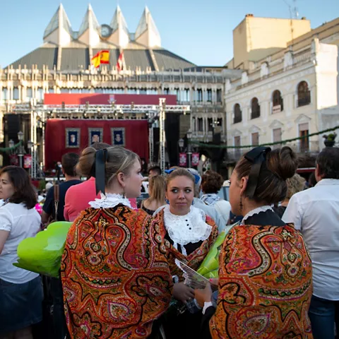 Grupo de mujeres con trajes tradicionales observando un escenario en una plaza llena de gente.