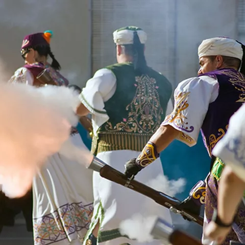 Participantes con trajes tradicionales disparando arcabuces durante un desfile.