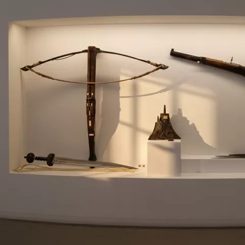 Nicho iluminado con ballesta, arma de fuego antigua y accesorios sobre pedestales.