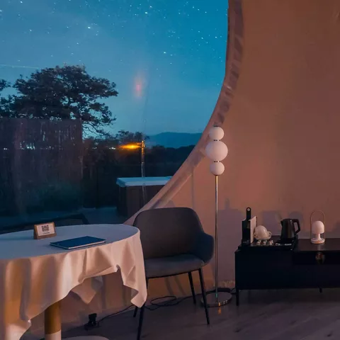 Interior de suite con cama, mesa y ventanal curvo transparente.