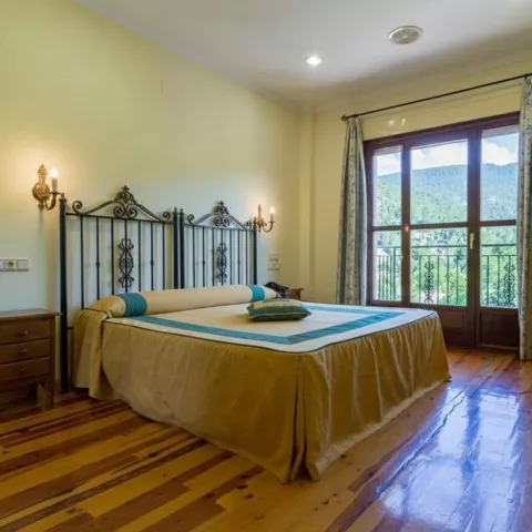 Habitación con cama doble, suelo de madera y ventanal con cortinas hacia el paisaje.