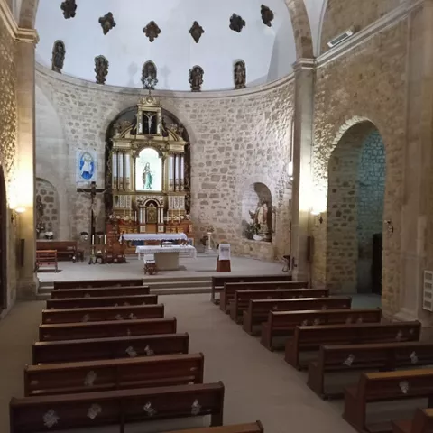 Interior de iglesia con bancos de madera y altar ornamentado frente a un ábside de piedra.