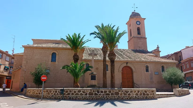 Fachada de iglesia con torre y palmeras en una plaza urbana