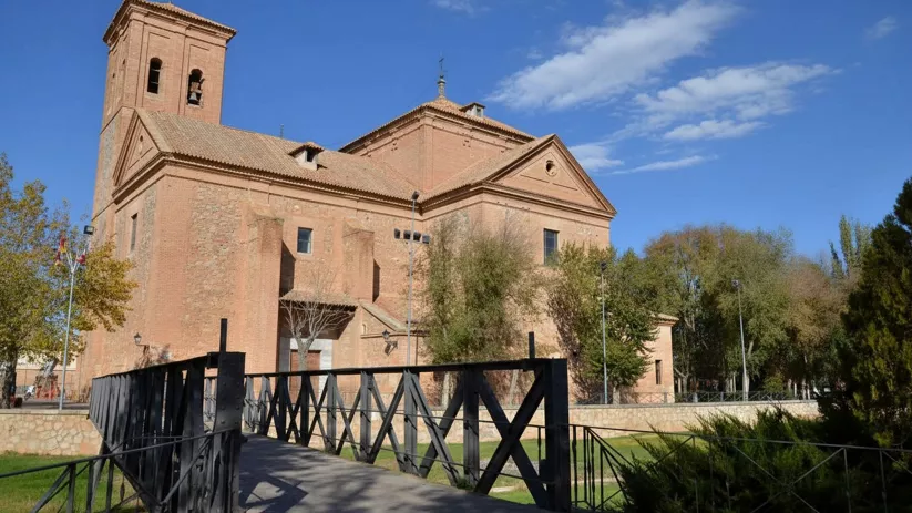 Iglesia de ladrillo junto a un puente metálico y zona arbolada en un día soleado.