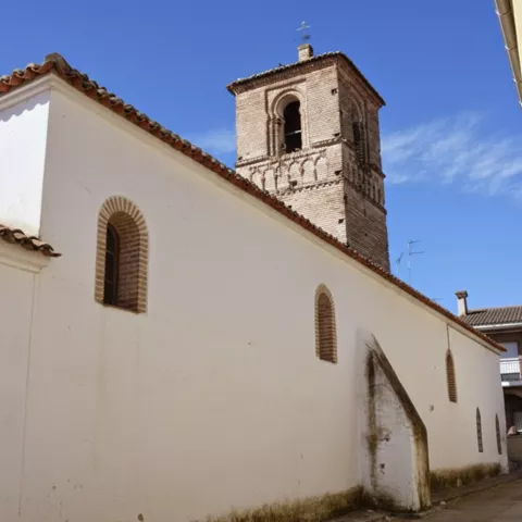 Perspectiva lateral de una iglesia blanca con pequeñas ventanas de arco, mostrando el contraste entre el muro liso y la torre de ladrillo al fondo.