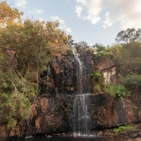 Cascada alta sobre pared de roca oscura, con poza tranquila y cielo claro.