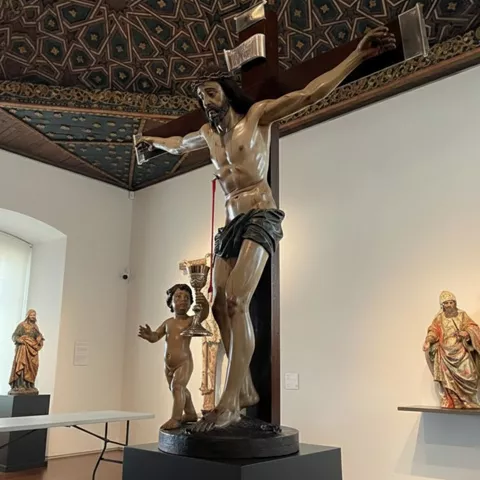 Escultura de Cristo en cruz expuesta en sala de museo con otras tallas religiosas al fondo.