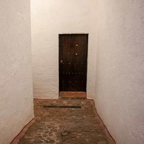 Pasillo estrecho encalado con suelo de piedra y puerta al fondo.