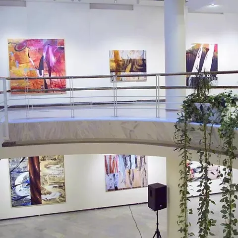 Galería interior con pinturas contemporáneas colgadas en paredes blancas y pasarela superior con plantas colgantes.