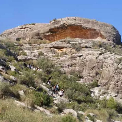 Abrigo rocoso elevado con sendero y personas caminando entre vegetación baja.