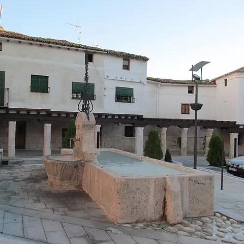 Plaza porticada con fuente de piedra y edificios blancos.