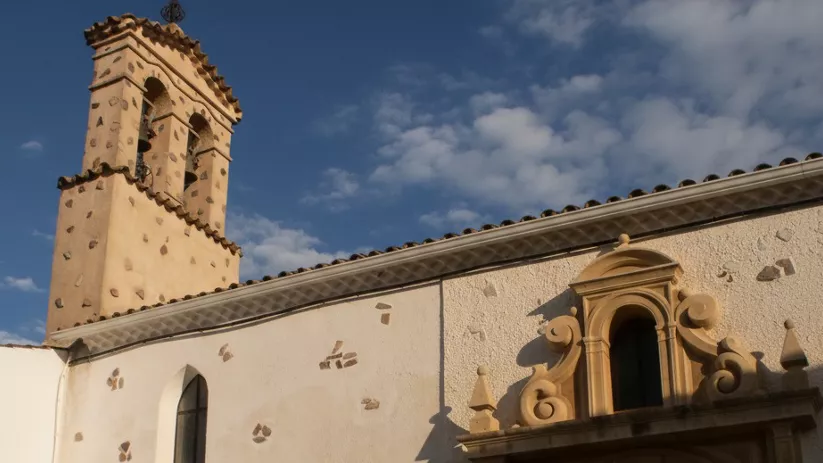 Iglesia de Povedilla con espadaña y portada barroca en una plaza soleada.