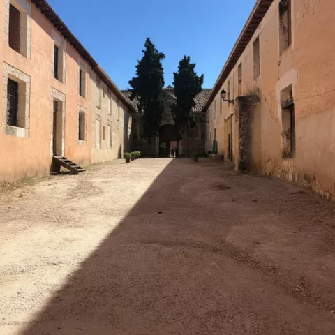 Patio alargado entre edificios antiguos con suelo de tierra y cipreses al fondo.