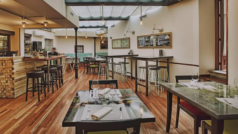 Zona de barra con taburetes altos, mesas altas y lámparas colgantes sobre suelo de madera.