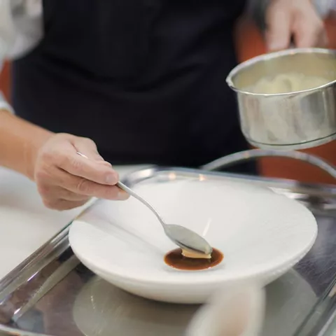 Cocinero emplatando salsa en un plato hondo sobre una bandeja metálica.