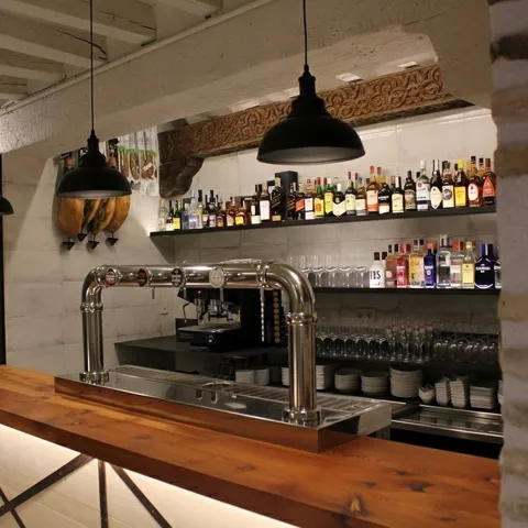 Barra de bar con grifos de cerveza, estanterías con botellas y lámparas colgantes negras.