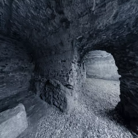 Interior de galería subterránea de piedra con arco y suelo de grava.